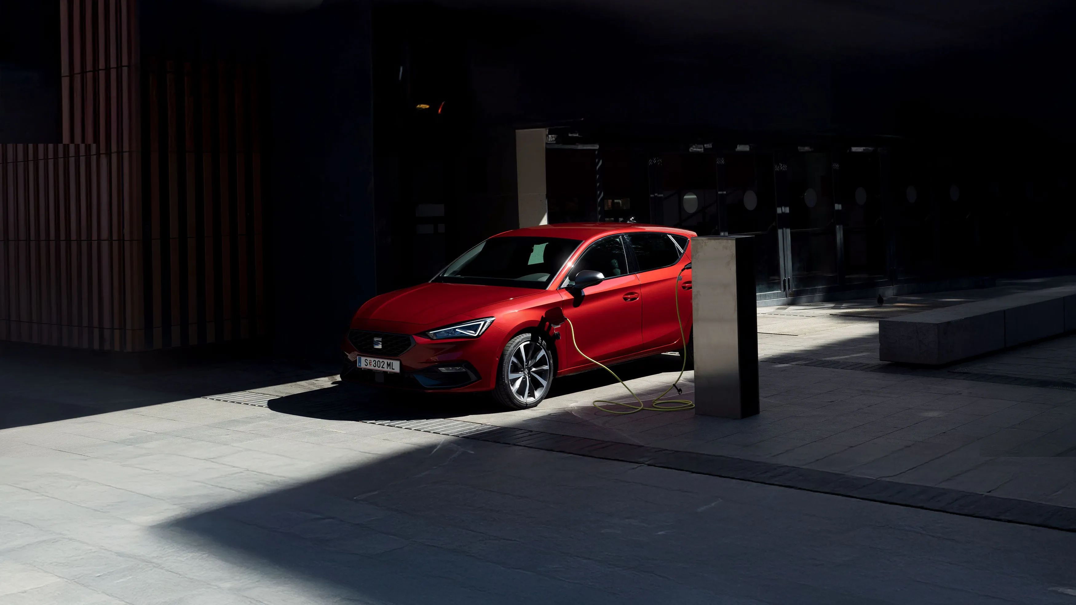 Vista lateral de um automóvel híbrido plug-in SEAT, na cor vermelha, estacionado e a carregar num espaço semiaberto com luz solar parcial, realçando a possibilidade de conduzir de forma eficiente, respeitando o ambiente e reduzindo as emissões.