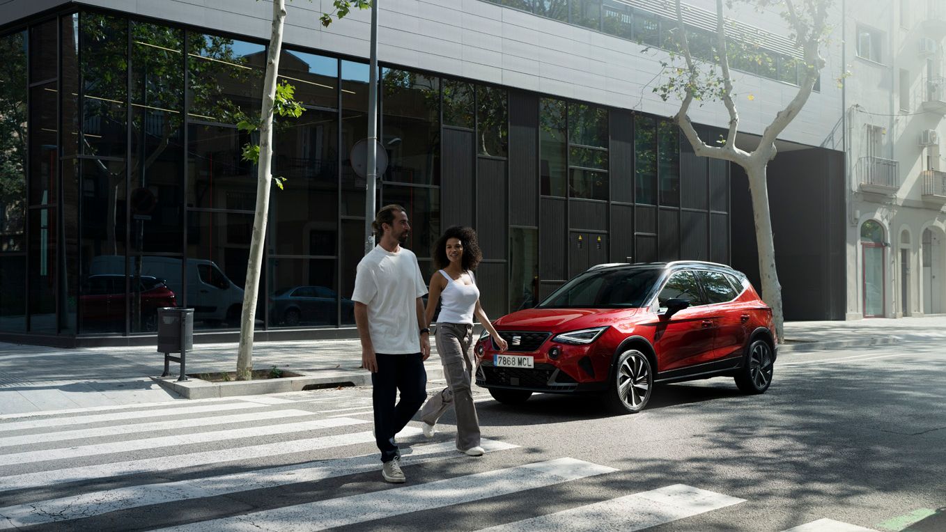 Homem e mulher atravessam a rua juntos, com um SEAT Arona na cor vermelho Desire estacionado atrás deles.