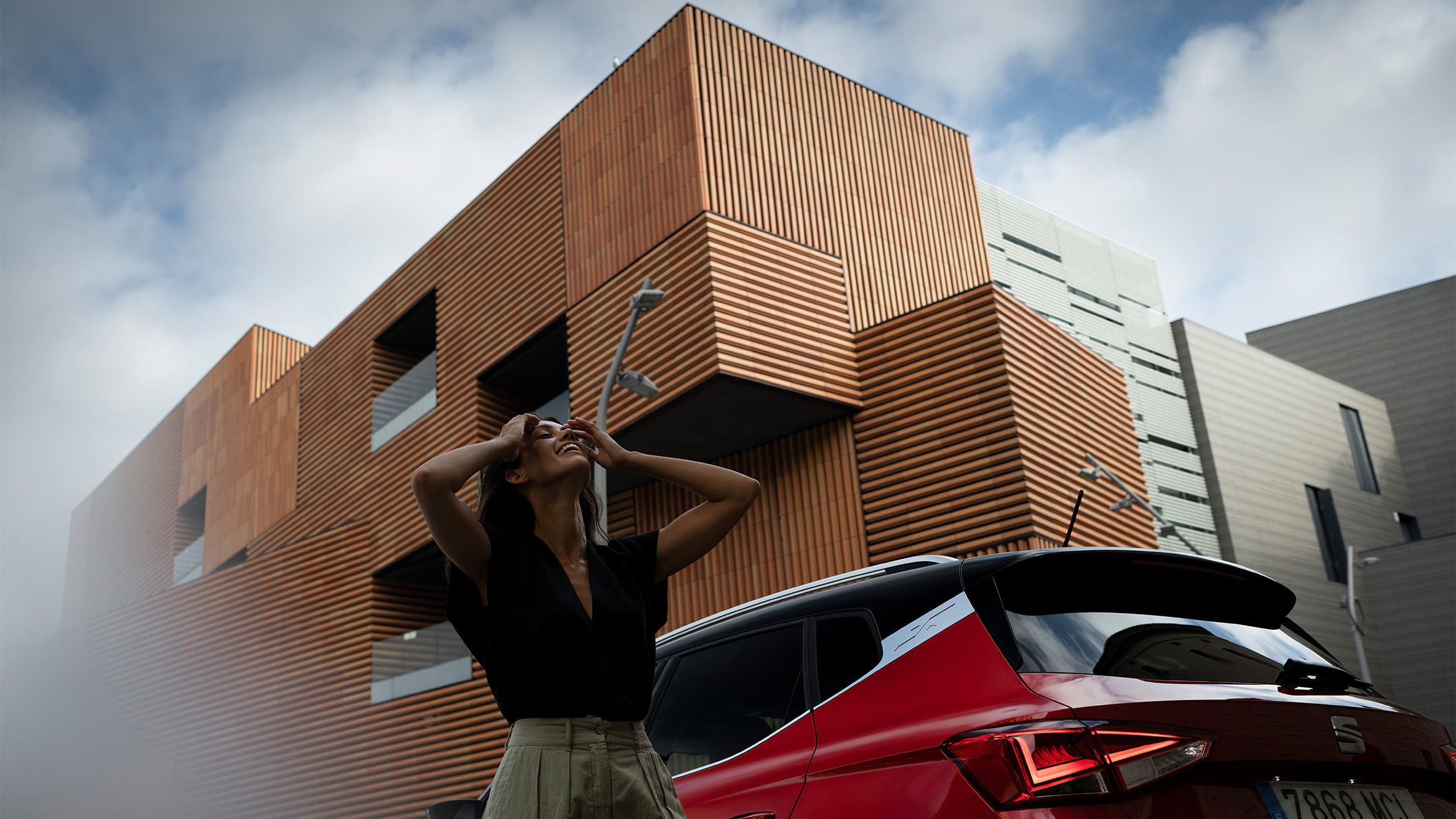 Mulher a olhar para cima e a sorrir, ao lado de um SEAT Arona na cor vermelho Desire, estacionado na rua